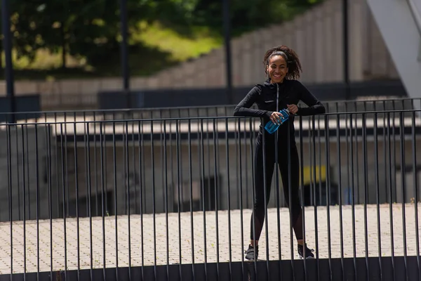 Deportiva afroamericana positiva sosteniendo botella deportiva y mirando la cámara en el puente - foto de stock