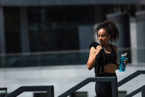 Deportista afroamericana sosteniendo chaqueta deportiva y botella cerca de barandilla al aire libre - foto de stock