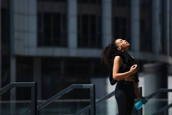 Seitenansicht einer afrikanisch-amerikanischen Sportlerin mit geschlossenen Augen, die Sportflasche und Jacke im Freien hält — Stockfoto