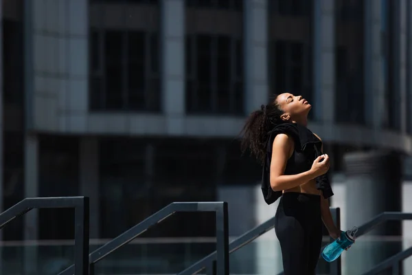 Side view of african american sportswoman with closed eyes holding sports bottle and jacket outdoors — Stock Photo