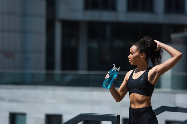 Vista lateral de una deportista afroamericana sosteniendo una botella deportiva y ajustando el cabello al aire libre — Stock Photo