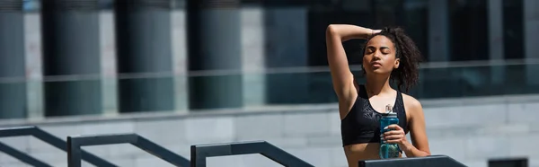 Junge afrikanisch-amerikanische Sportlerin mit geschlossenen Augen hält Sportflasche auf städtischer Straße nahe Geländer, Banner — Stockfoto