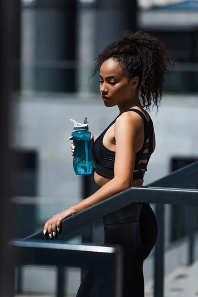 Bonita y en forma deportista americana afican sosteniendo la botella de los deportes mientras que está parado cerca de barandilla - foto de stock