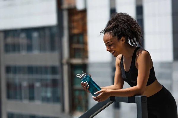 Vue latérale d'une sportive afro-américaine heureuse regardant une bouteille de sport dans une rue urbaine — Photo de stock