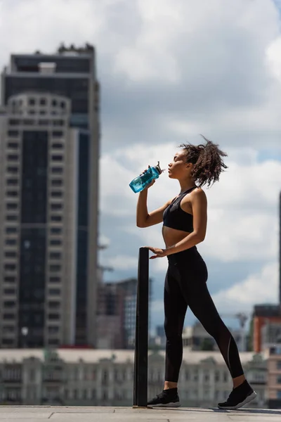 Vista laterale della sportiva afro-americana che beve acqua dalla bottiglia sportiva sulla strada urbana — Foto stock