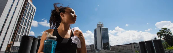Низкий угол обзора африканской спортсменки с полотенцем и спортивной бутылкой на открытом воздухе, баннер — стоковое фото