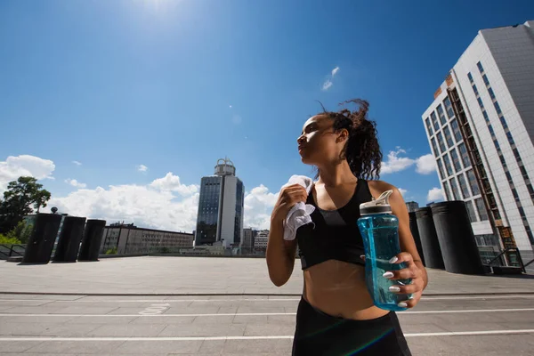 Side view of african american sportswoman holding towel and sports bottle on urban street — Stock Photo