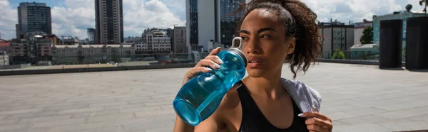Deportiva afroamericana bebiendo agua y sosteniendo toalla en la calle urbana, pancarta - foto de stock
