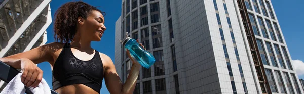 Low angle view of smiling african american sportswoman with towel holding sports bottle on urban street, banner — Stock Photo