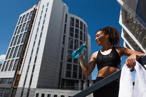 Visão de baixo ângulo da esportista afro-americana positiva com toalha segurando garrafa de esportes na rua urbana — Fotografia de Stock