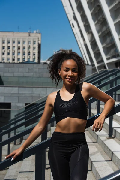 Femme sportive afro-américaine souriante regardant une caméra près d'un escalier avec une rampe dans une rue urbaine — Photo de stock