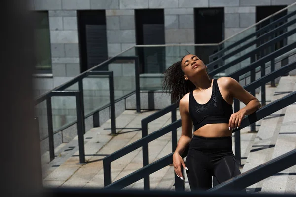 Pretty african american sportswoman with closed eyes standing on stairs near railing — Stock Photo