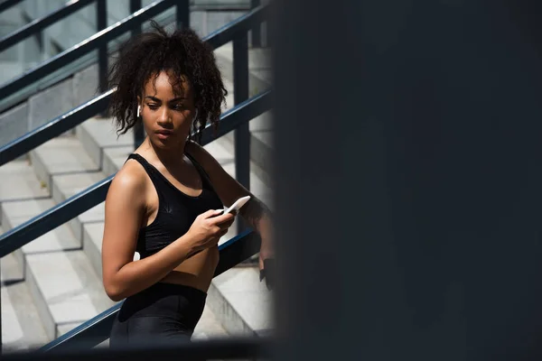 Deportista afroamericana en auricular sosteniendo smartphone y mirando hacia otro lado en las escaleras - foto de stock