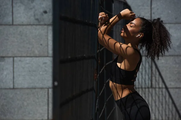 Side view of african american sportswoman in earphone standing near fence outdoors — Stock Photo