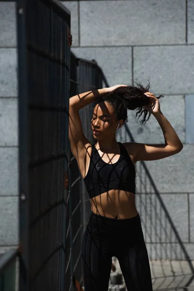 African american sportswoman in earphone adjusting hair near fence — Stock Photo