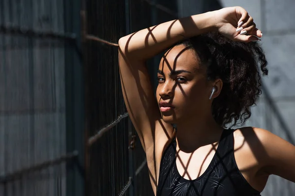 Pretty african american sportswoman in wireless earphone looking at camera near fence — Stock Photo