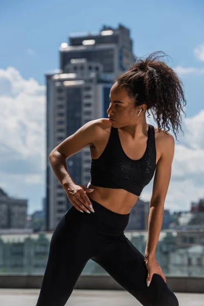 Deportista afroamericana en ropa deportiva negra y auricular mirando hacia otro lado en la calle urbana - foto de stock