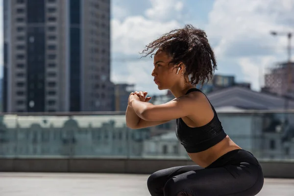 Side view of african american sportswoman in earphone doing squat on urban street — Stock Photo