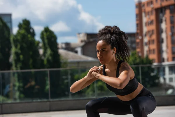 African american sportswoman in earphone doing squat while training outdoors — Stock Photo