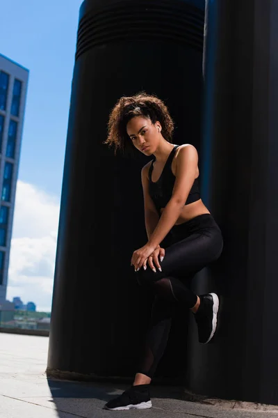 Deportista afroamericana en auricular mirando la cámara cerca del edificio - foto de stock