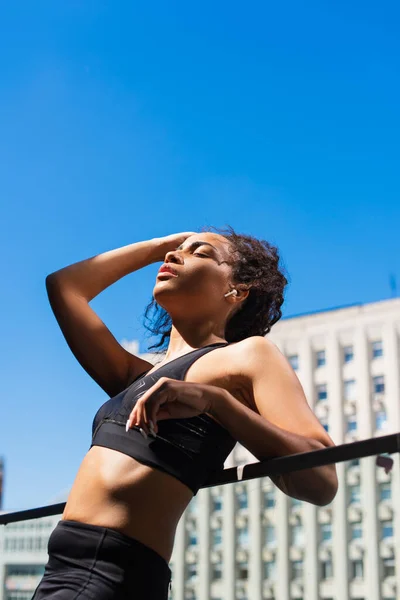 Ajuste afroamericana deportista en auriculares descansando en la calle urbana con el cielo en el fondo - foto de stock