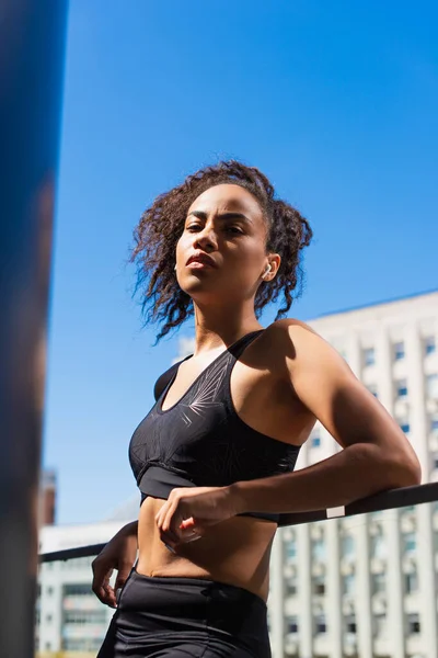 Femme afro-américaine en écouteur et vêtements de sport debout dans la rue urbaine le jour — Photo de stock