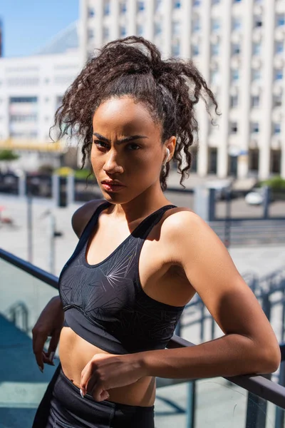 Confident african american sportswoman in wireless earphone looking at camera near railing on urban street — Stock Photo