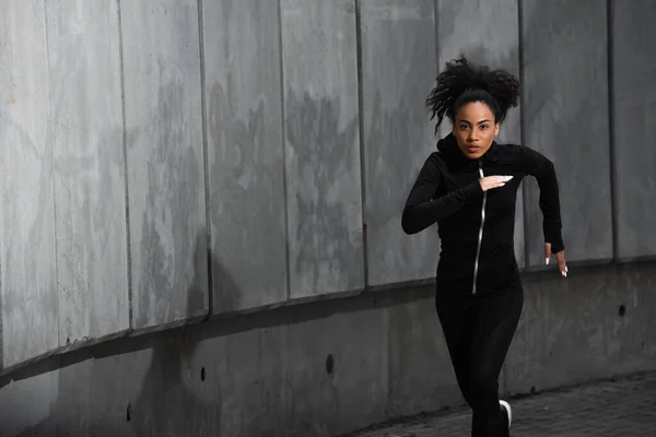 Focada mulher afro-americana em preto jaqueta de esportes correndo ao ar livre — Fotografia de Stock