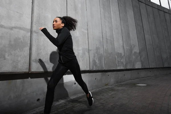 Joven deportista afroamericana corriendo cerca de muro de hormigón con sombra en calle urbana - foto de stock