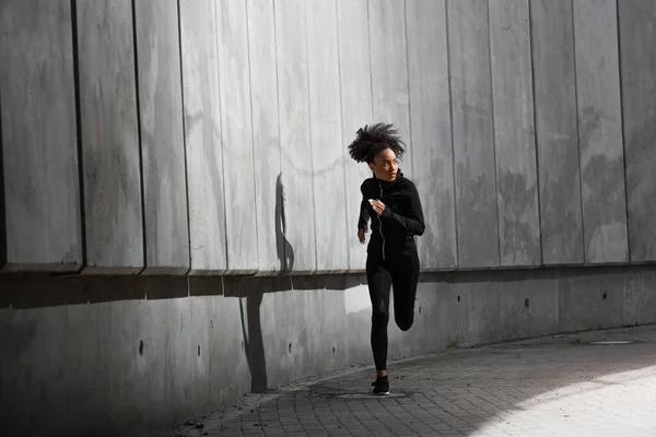 Young african american woman running while training near building with concrete wall — Stock Photo