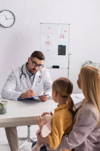 Pediatrician with clipboard looking at patient and parent in hospital — Stock Photo