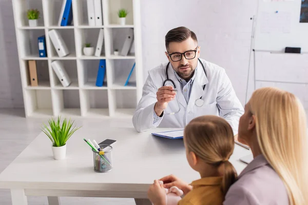 Pediatrician in eyeglasses looking at mother and kid in clinic — Stock Photo