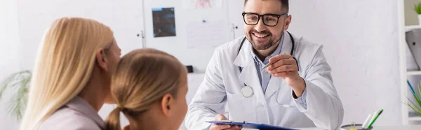 Smiling doctor with clipboard pointing at mother with child in clinic, banner — Stock Photo