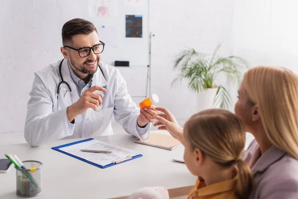 Médico sonriente sosteniendo frasco con pastillas cerca de la madre y el niño - foto de stock