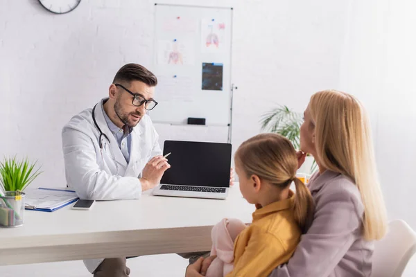 Family doctor pointing at laptop near mother and daughter in clinic — Stock Photo
