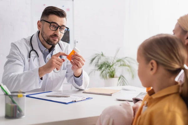 Доктор в окулярах вказує на банку з таблетками поблизу розмитої матері і дитини — стокове фото