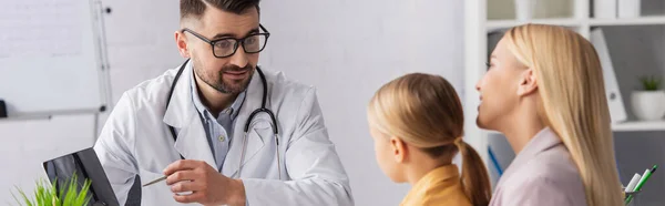 Médico mirando al niño mientras apunta a la computadora portátil cerca de los padres sonrientes, pancarta - foto de stock