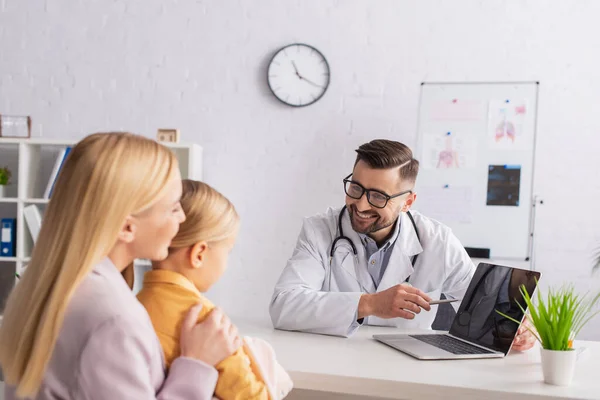 Medico di famiglia positivo che punta al computer portatile con schermo vuoto vicino a madre e bambino in ospedale — Foto stock