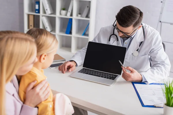 Doctor in eyeglasses pointing at laptop with blank screen near clipboard and family in hospital — Stock Photo