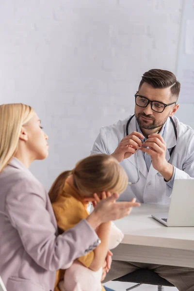 Family doctor looking at blurred mother talking near kid in clinic — Stock Photo