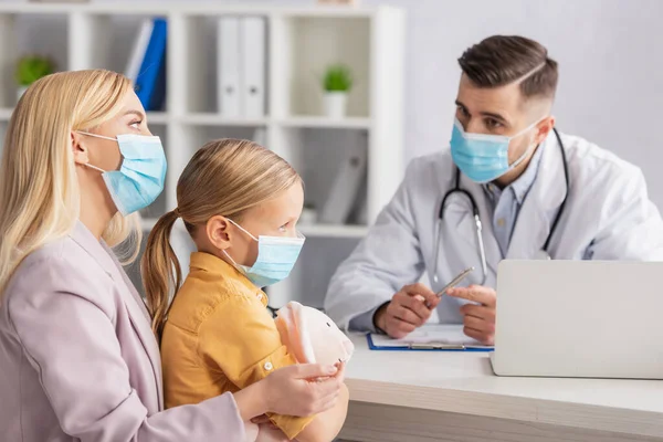 Мати і дитина в медичних масках, що сидять біля розмитого лікаря і ноутбука — стокове фото