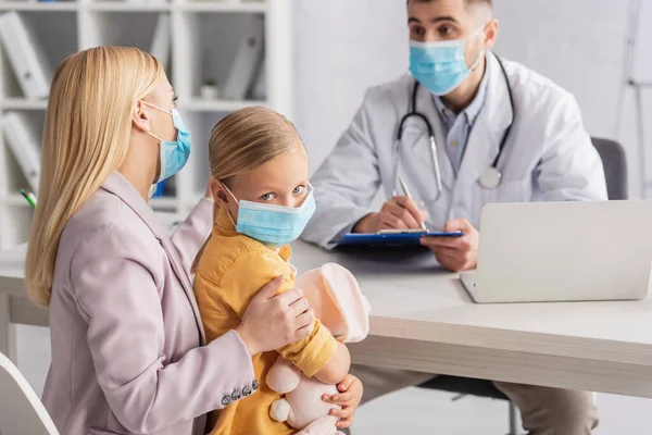 Niño en máscara médica mirando a la cámara cerca de la madre y el médico de familia borrosa - foto de stock