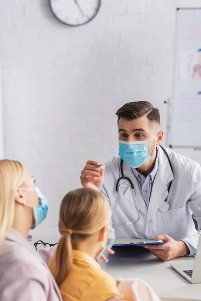 Medico in maschera medica che tiene appunti e penna vicino a madre e bambino — Foto stock