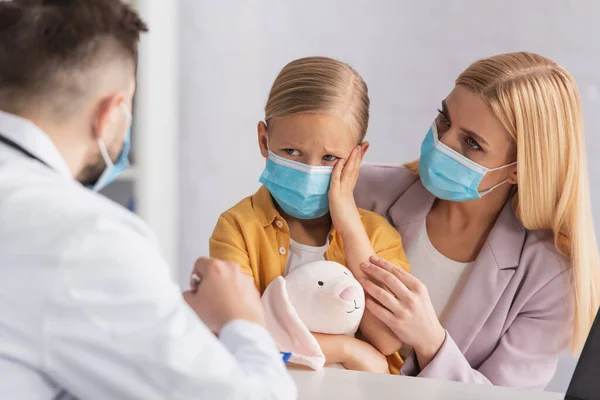 Kid touching head near mother and blurred doctor in white coat — Stock Photo