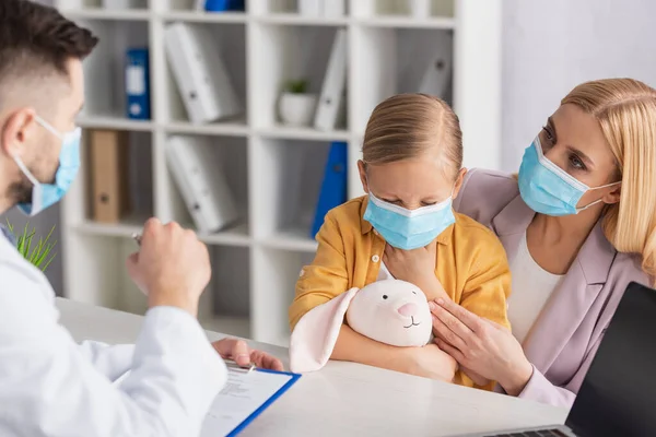 Mother in medical mask looking at doctor with clipboard near diseased child — Stock Photo