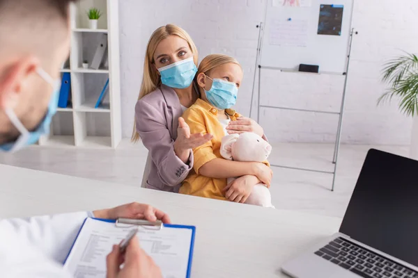 Mother in medical mask pointing at pediatrician near child and laptop — Stock Photo