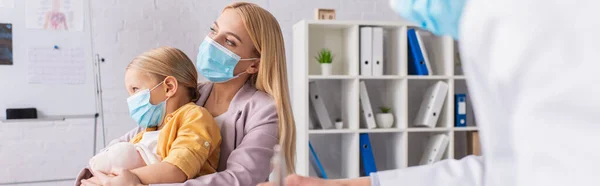 Madre en máscara médica abrazando a la hija con juguete suave cerca del médico borroso en la clínica, pancarta - foto de stock