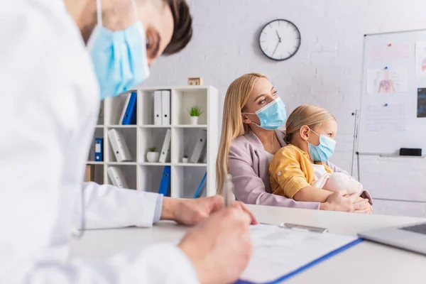 Woman in medical mask hugging child near family doctor writing on clipboard in clinic — Stock Photo