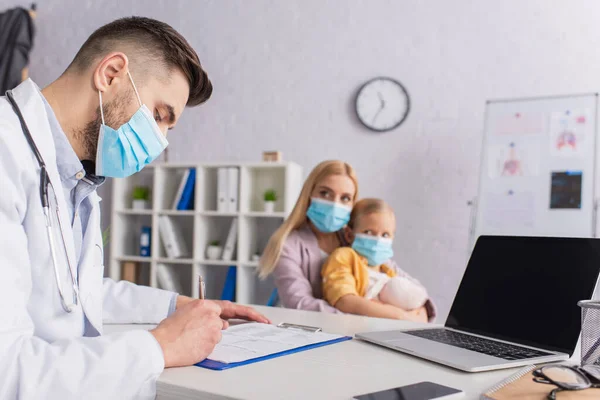 Врач в медицинской маске пишет на планшете рядом с устройствами и семьей в клинике — стоковое фото