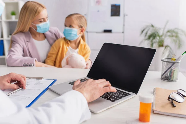 Doctor using laptop near pills and mother with daughter in medical masks on blurred background — Stock Photo